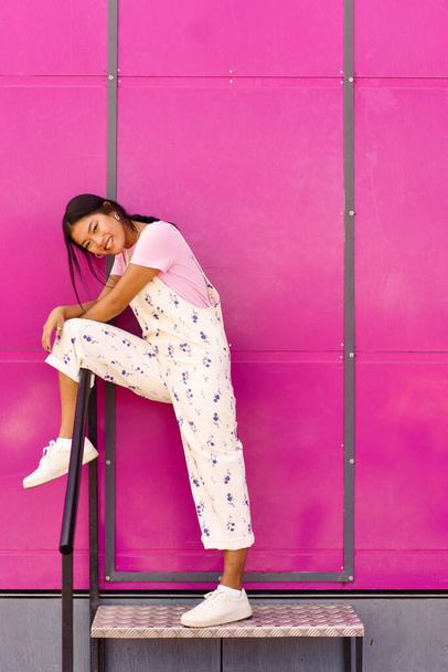 Corps complet jeune femme asiatique heureuse, dans des vêtements élégants tenant la jambe sur la rampe et regardant la caméra avec le sourire tout en se tenant debout sur le pas en métal contre un mur rose vif - Photo, image