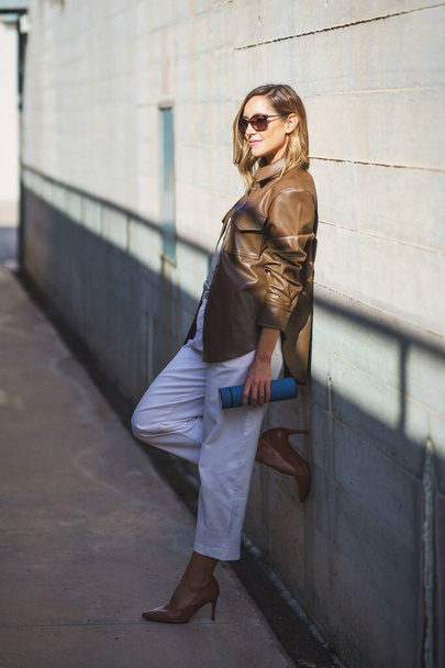 Full body woman in stylish outfit and sunglasses with thermos leaning on sunlit building wall in daytime on city street - Photo, Image
