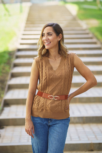 Pozitivní dospělá žena ve stylovém pleteném top drží ruku v pase a dívá se pryč s úsměvem na rozmazaném pozadí schodů - Fotografie, Obrázek