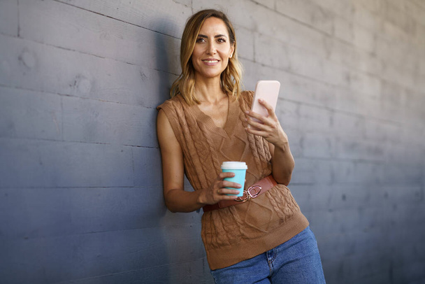 Γυναίκα σε κομψό πλεκτό τοπ, με φλιτζάνι καφέ για να πάει κοιτάζοντας μακριά με χαμόγελο και τη χρήση smartphone, ενώ ακουμπά σε γκρι τοίχο στο δρόμο της πόλης - Φωτογραφία, εικόνα