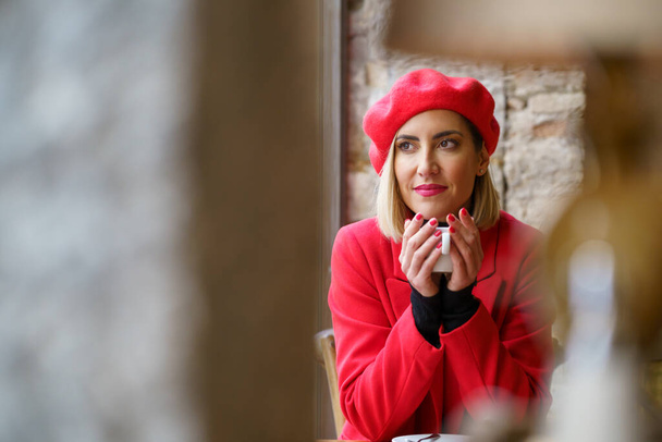 Через размытую колонну и лампу очаровательной женщины в красной одежде, смотрящей в сторону, сидя с чашкой горячего напитка в современном кафетерии - Фото, изображение