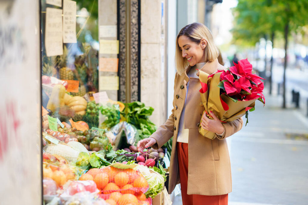 Zijaanzicht van glimlachende vrouw met boeket bloemen kopen van verse groenten terwijl staan op stal op straat van de stad - Foto, afbeelding