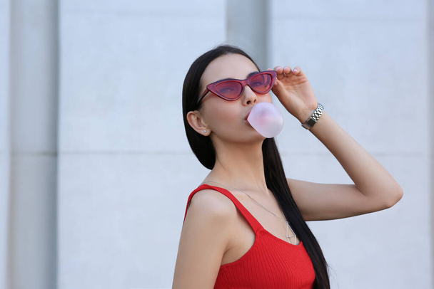 Beautiful woman in stylish sunglasses blowing gum near wall outdoors - Fotografie, Obrázek
