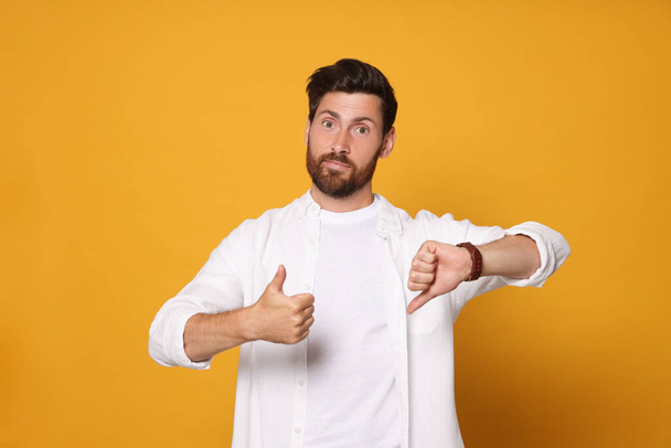 Handsome bearded man showing thumbs up and down on orange background - Фото, зображення