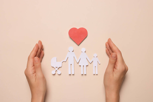 Woman protecting paper family figures and red heart on beige background, top view. Insurance concept - Foto, Imagem
