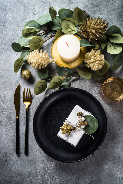 Christmas table setting with black plate, candles, eucalyptus leaves and golden decorations. Top view at stone table. - Photo, image