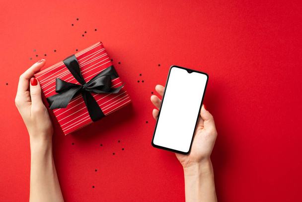 Black friday online shopping concept. First person top view photo of female hands holding smartphone and big red giftbox with ribbon bow over confetti on isolated red background with blank space - Foto, Imagen