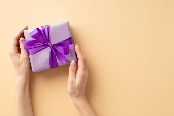 New Year concept. First person top view photo of female hands holding lilac giftbox with violet ribbon bow on isolated pastel beige background with empty space - Foto, imagen