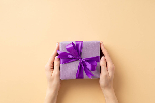 Concept du Nouvel An. Première personne vue du dessus photo de mains féminines donnant boîte-cadeau pourpre avec ruban violet arc sur fond beige pastel isolé avec espace vide - Photo, image