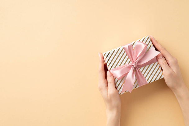 New Year concept. First person top view photo of woman's hands giving present box with pink ribbon bow on isolated pastel beige background with blank space - Fotoğraf, Görsel