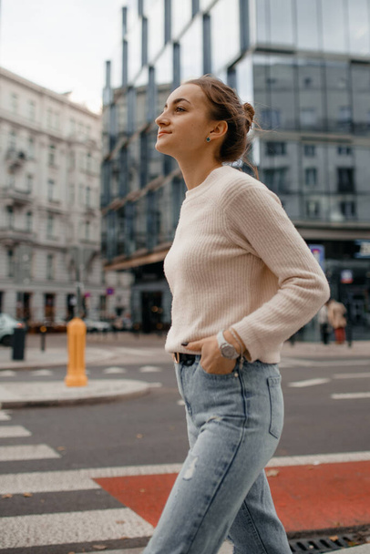 Cool girl in sweater looks away. Lifestyle portrait of a young stylish woman walking outdoors. Lifestyle portrait of a young woman in sweater standing on the street in Warsaw. - Valokuva, kuva