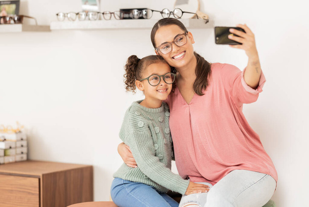 Mutter, Mädchen und Handy-Selfie beim Augenarzt, Brille und Lächeln für das Augenlicht, Sehvermögen und Social-Media-Bild für den neuen Linsenrahmen. Frau, Kind und glücklich mit Brille oder 5g Handy. - Foto, Bild