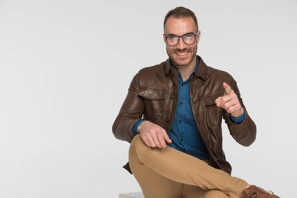 sexy casual guy pointing at the camera while sitting on a chair and wearing eyeglasses - Фото, изображение