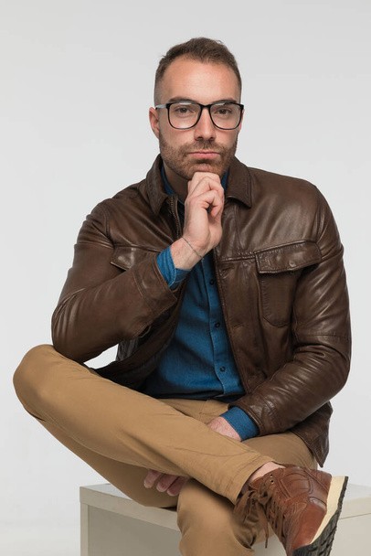coole lässige Mann sitzt auf einem Stuhl, während seine Beine kreuzen und trägt eine braune Lederjacke und Brille auf grauem Studiohintergrund - Foto, Bild