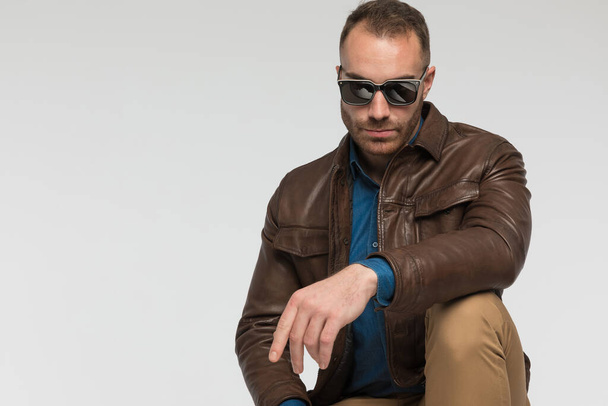 sexy casual man is posing with tough attitude and wearing a brown leather jacket and sunglasses on gray studio background - Foto, Imagem