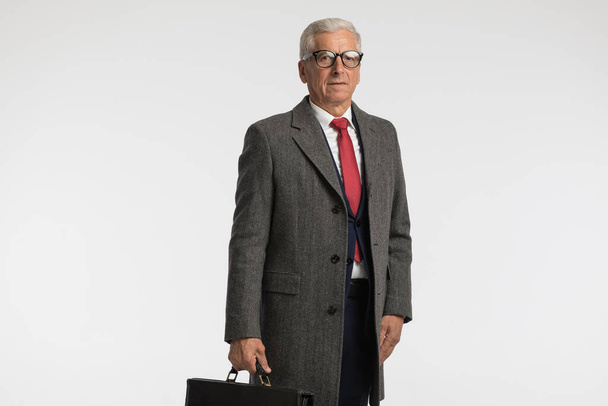 El hombre de negocios con el pelo canoso está firme, vistiendo un abrigo gris y sosteniendo un maletín contra el fondo gris del estudio. - Foto, Imagen