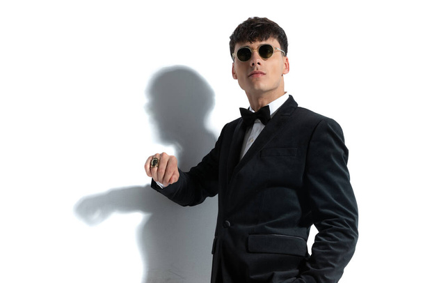 Young businessman is showing off his ring, putting one hand in pocket, and wearing sunglasses against white studio background - Photo, Image