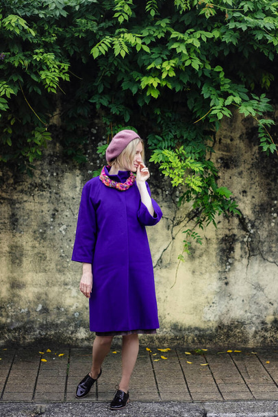 A woman in a coat and beret poses by a wall covered with greenery. - Fotografie, Obrázek
