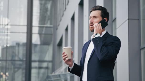 Young man standing outdoors talking on mobile phone holds paper cup with coffee professional businessman answering business call communicate with client male customer orders delivery using smartphone - Footage, Video