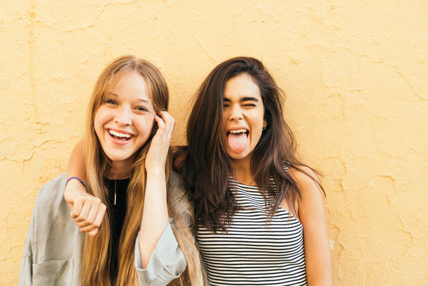 One brunette and one blonde couple of friends standing against a yellow wall outdoors sticking tongue out and having a good time - Фото, зображення