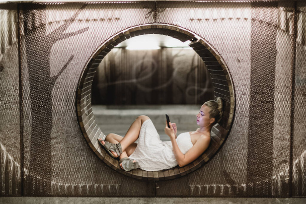 Дама в білій літній сукні грає недбало зі своїм смартфоном андроїд, переглядаючи нерівні соціальні мережі, що розслабляються на сучасній круговій лавці в міському пішохідному проході
. - Фото, зображення