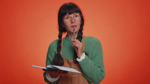 Στοχαστική δημοσιογράφος γυναίκα με πράσινο πουλόβερ κάνοντας σημειώσεις, γράφοντας τις σκέψεις με στυλό σε σημειωματάριο ημερολόγιο, να κάνει λίστα, καλή ιδέα. Νεαρή ενήλικη κοπέλα απομονωμένη σε φόντο πορτοκαλί στούντιο - Πλάνα, βίντεο