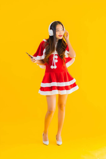 Portrait belle jeune asiatique vêtements de Noël et chapeau écouter de la musique avec casque et smartphone sur fond isolé jaune
 - Photo, image