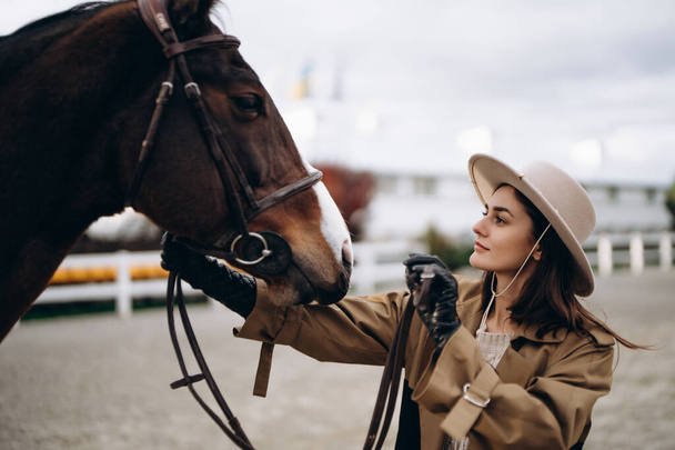 A young woman in a brown coat and hat near a horse. High quality photo - Foto, Imagen