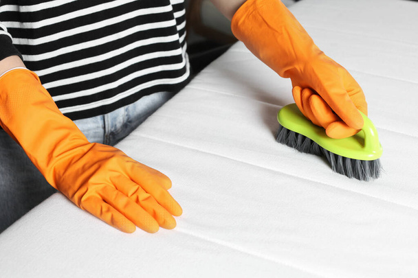 Woman in orange gloves cleaning white mattress with brush indoors, closeup - Фото, изображение
