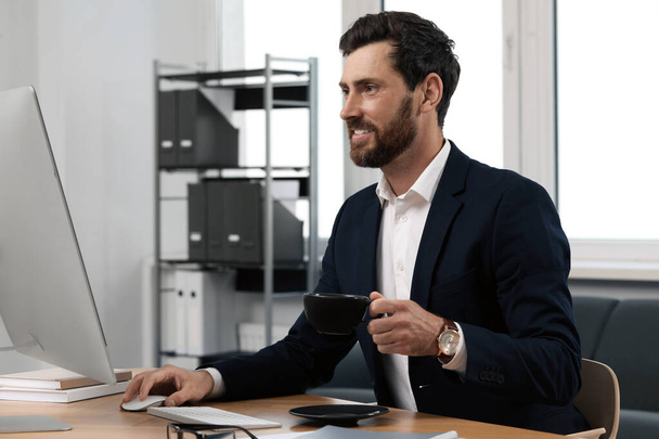 Handsome bearded man with cup of drink working on computer at table in office - Foto, immagini