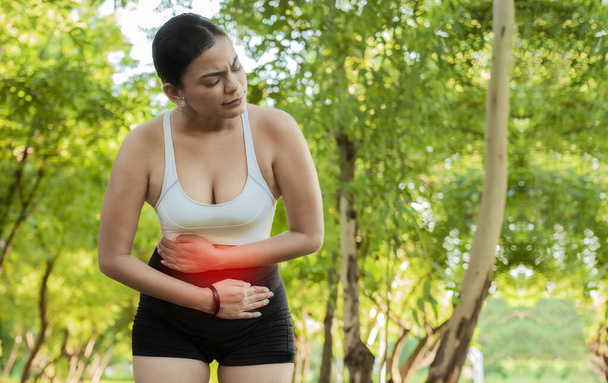 Woman in sportswear with stomach ache in a park. Running woman with digestive problems. Sport girl with stomach ache outdoors with copy space. Fatigued young woman with stomach ache - Photo, Image