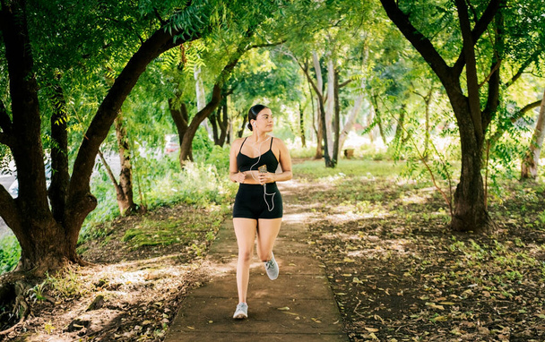 Lifestyle of sporty young woman running in a park surrounded by trees. Healthy lifestyle concept, Sporty young woman running in a park. A girl running in a park while listening to music - Фото, зображення