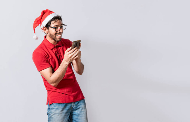 Glimlachende jongeman met kerstmuts met geïsoleerde mobiele telefoon. Een gelukkige man met een kerstmuts die lacht naar een geïsoleerde mobiele telefoon. Mensen in de kerstman hoed met behulp van en glimlachen naar mobiele telefoon geïsoleerd - Foto, afbeelding
