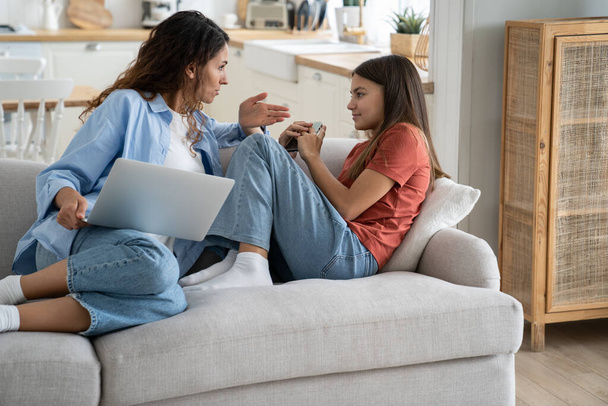Displeased casual European woman scolding teenage daughter for using mobile phone for too long or being addicted to apps. Young mother with laptop sits on sofa talking to discouraged school age girl - Фото, изображение