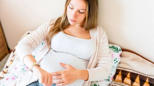 Těhotné bolestivé kontrakce. Těhotná žena se dívá na hodiny, drží malé břicho. Čas porodu, stahy bolesti. Těhotenství, medicína koncepce zdravotní péče - Fotografie, Obrázek