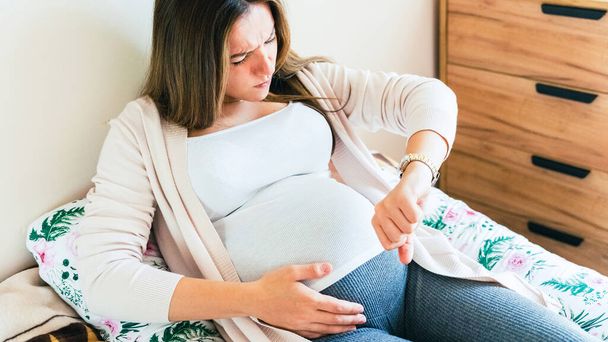 Έγκυες συσπάσεις πόνου. Έγκυος γυναίκα παρακολουθεί ρολόι, κρατάει την κοιλιά του μωρού. Ώρα τοκετού, πόνος στις συσπάσεις. Εγκυμοσύνη, ιατρική υγειονομική περίθαλψη έννοια - Φωτογραφία, εικόνα