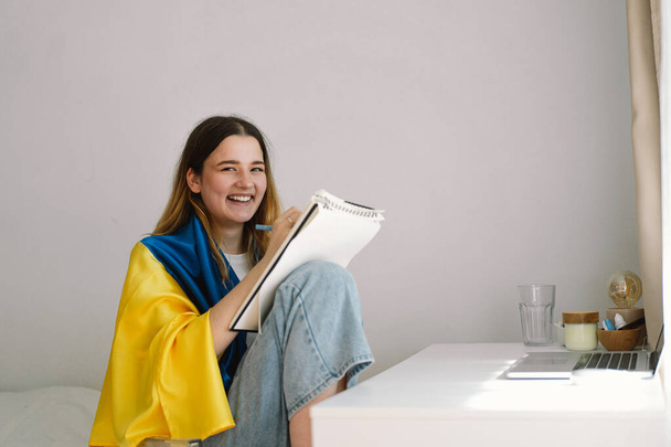 Una adolescente ucraniana está envuelta en la bandera ucraniana y está dibujando o haciendo tareas en el dormitorio. Un adolescente estudia en casa. El concepto de estudios durante la guerra en Ucrania. - Foto, imagen