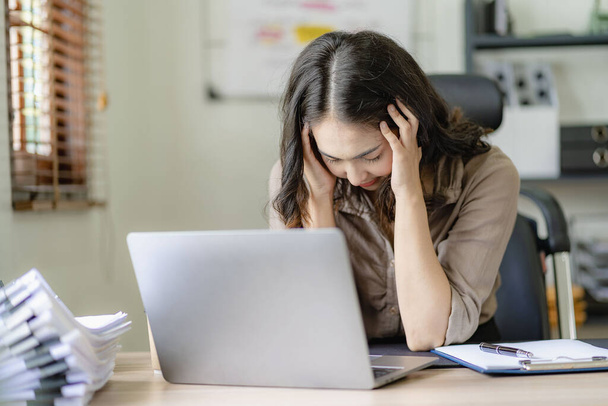 Asiática mujer de negocios dolor de cabeza durante el trabajo análisis de gráficos financieros con portátil en su escritorio mujer estresada Dolor de cabeza durante las horas extras en la oficina en casa - Foto, imagen
