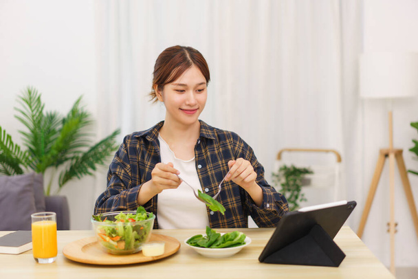 Стиль жизни в гостиной концепции, молодая азиатская женщина смотрит на планшет и едят овощной салат. - Фото, изображение
