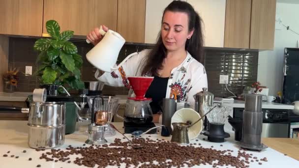 Siyah saçlı güzel genç bir kadın kahve yapmak için çeşitli aksesuarlar ve kahve çiçeğinin yanındaki masaya farklı profesyonel kahve ekipmanları döker. - Video, Çekim