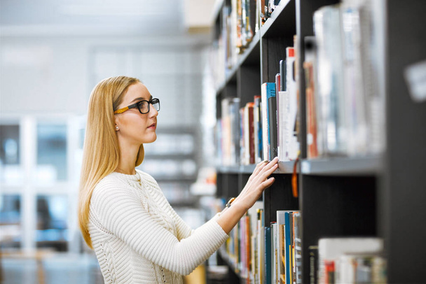 Tantos para escolher. uma jovem focada pegando um livro para ler de uma estante dentro de uma biblioteca - Foto, Imagem