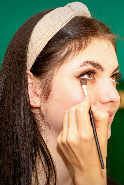 Maquillage dans le processus. La main du maquilleur applique une ombre à paupières sous les yeux d'une jeune belle femme modèle caucasienne - Photo, image