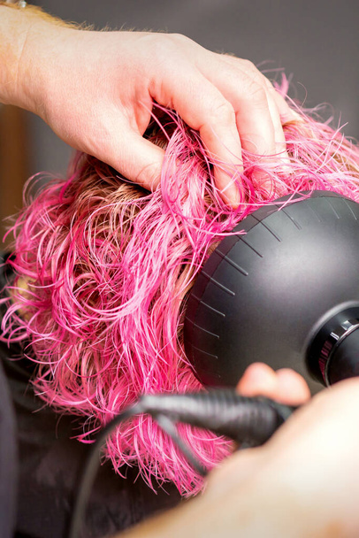 Asciugatura di capelli corti rosa bob di una giovane donna caucasica con un asciugacapelli nero con la spazzola per mano di un parrucchiere maschile in un salone di parrucchiere, primo piano - Foto, immagini
