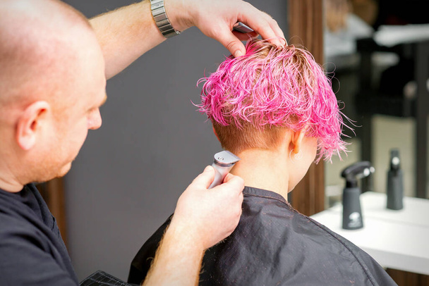 Cabeleireiro masculino rapa o pescoço de uma jovem mulher caucasiana com um penteado rosa curto por barbeador elétrico em um salão de cabeleireiro, close-up - Foto, Imagem