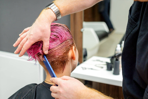 Парикмахер расчесывает окрашенные розовые влажные короткие волосы клиентки в парикмахерской, вид сзади - Фото, изображение