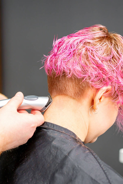 Male hairdresser shaves neck of a young caucasian woman with a short pink hairstyle by electric shaver in a hairdresser salon, close up - Photo, Image