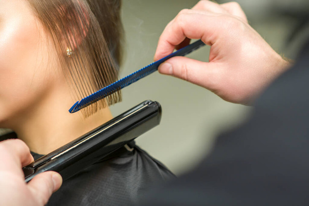 Hairstylist is rechttrekken kort haar van jonge brunette vrouw met een plat strijkijzer in een kapsalon, close-up - Foto, afbeelding