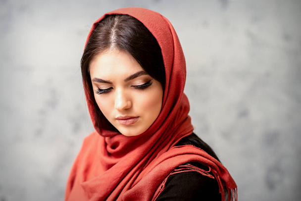 De modieuze jonge vrouw. Portret van het prachtige vrouwelijke model met lang haar en make-up met wimper verlengstukken in een rode sjaal. schoonheid jong Kaukasisch vrouw op de achtergrond van een grijze muur - Foto, afbeelding
