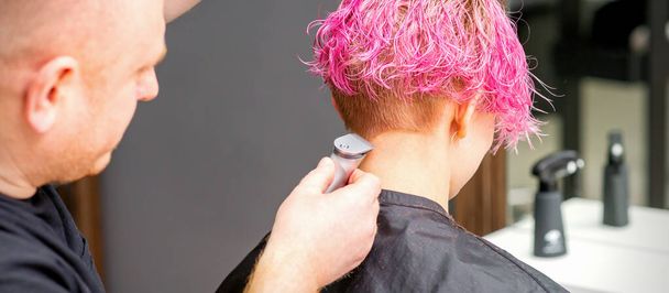 Mies kampaaja ajelee nuoren valkoihoisen naisen kaulan, jolla on lyhyt vaaleanpunainen kampaus sähköisellä parranajokoneella kampaamossa, lähikuva - Valokuva, kuva