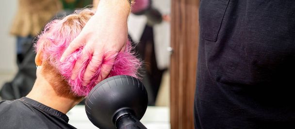 Secado corto peinado rosa bob de una joven mujer caucásica con un secador de pelo negro con el cepillo por las manos de un peluquero masculino en una peluquería, de cerca - Foto, imagen
